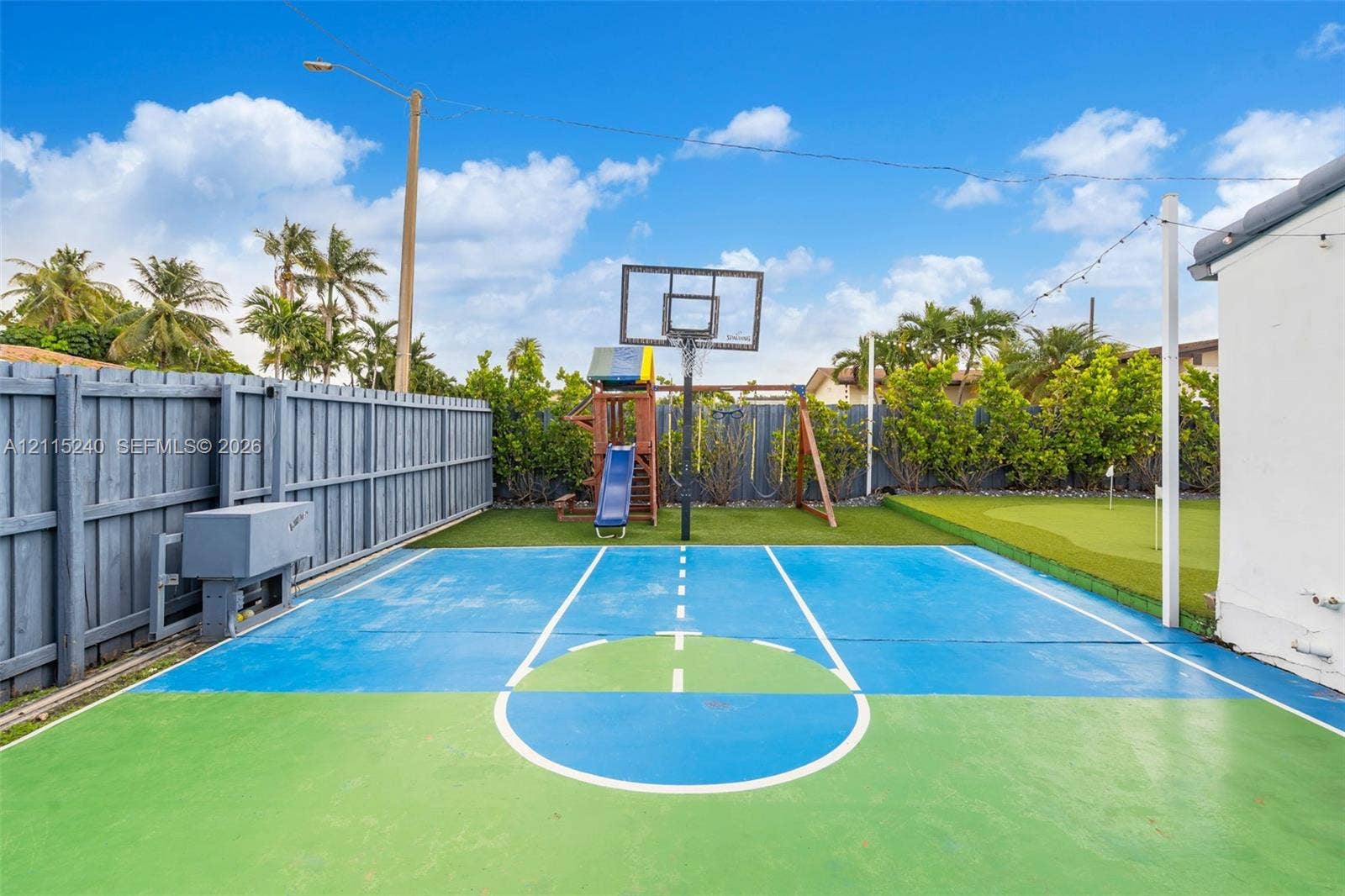 Outdoor area featuring a painted hard-surface court, a built-in basketball hoop, a dedicated putting green, and enclosed by wood-panel fencing