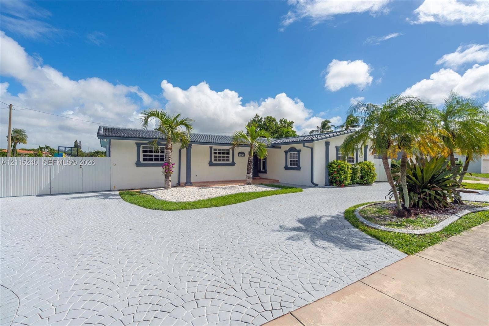 Single-story exterior featuring a decorative paved driveway, white stucco finish, dark shingle roof, integrated landscaping beds, and a white privacy fence
