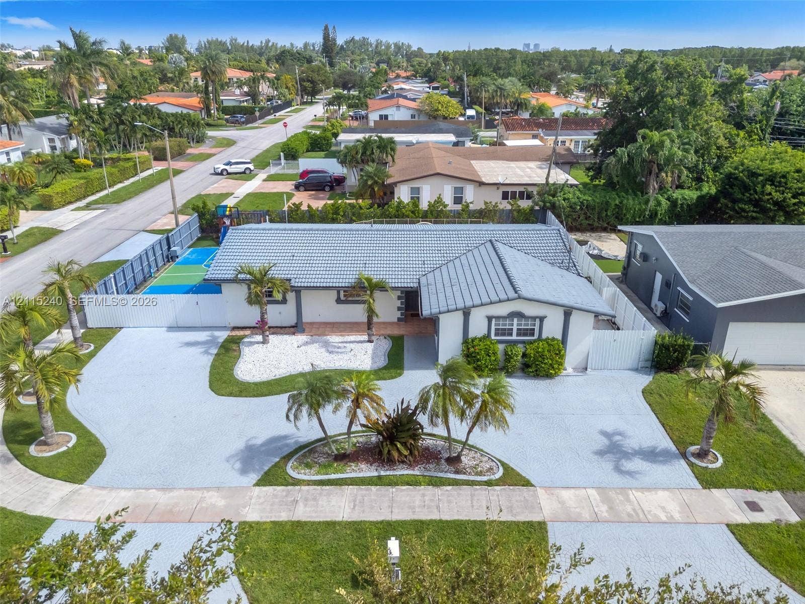 Single-story residence featuring a tile roof, an expansive paved driveway, a gated sports court, and manicured landscaping with mature palm trees