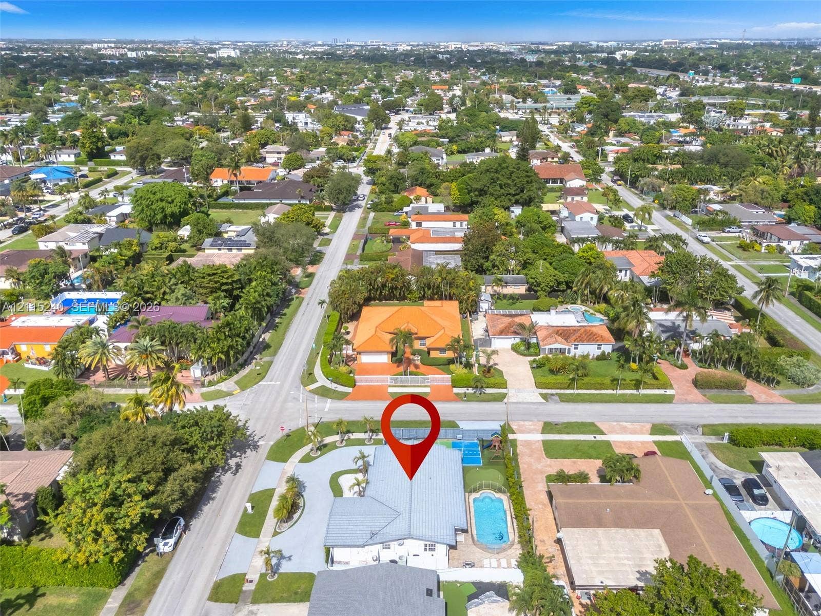 Aerial view of corner lot property featuring a light-toned multi-gabled roof, an expansive paved driveway, a private in-ground pool, and mature perimeter landscaping