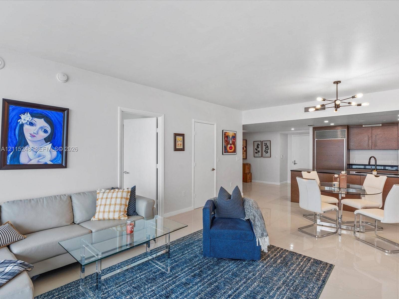 Open-concept interior featuring light-toned tile flooring, recessed ceiling lighting, a modern multi-arm chandelier, and a built-in cabinetry kitchen with an integrated refrigerator