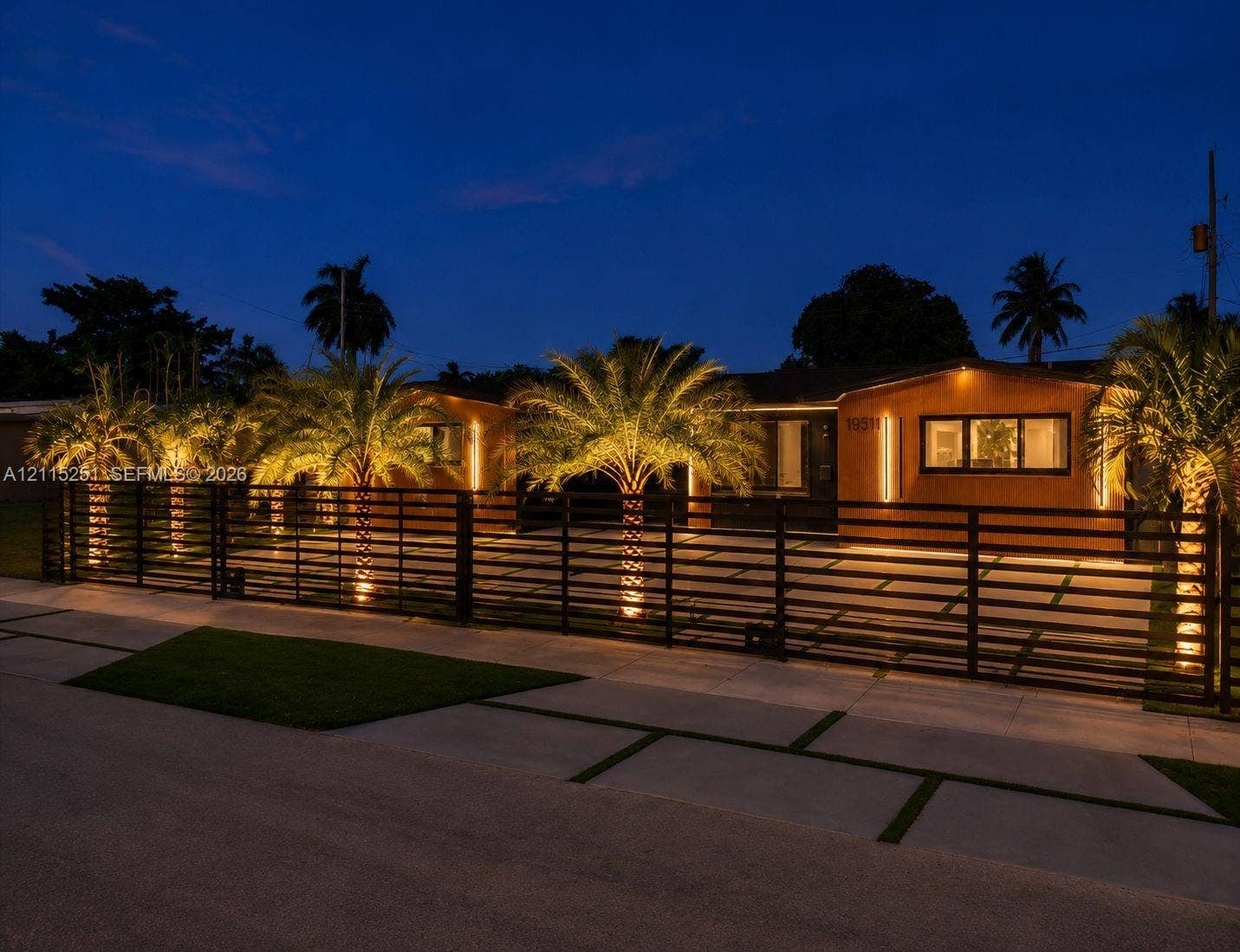 19511 NE 18th Place, Miami, FL 33179 Single-story residence featuring horizontal wood-finish siding, a wide metal horizontal slat perimeter fence, paved driveway with grass grid accents, and mature palm trees