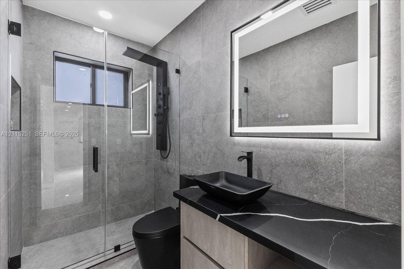 19511 NE 18th Place, Miami, FL 33179 Bathroom featuring full-height large-format wall tile, glass-enclosed shower with matte black rainfall panel, floating vanity with black countertop, rectangular vessel sink, and illuminated vanity mirror