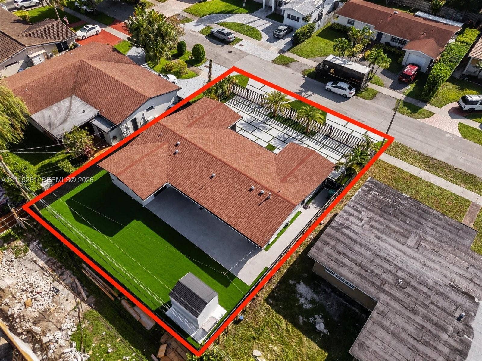 19511 NE 18th Place, Miami, FL 33179 Aerial perspective featuring a U-shaped roofline, paved driveway with grass grid detail, synthetic turf lawn, perimeter fencing, and mature palm trees
