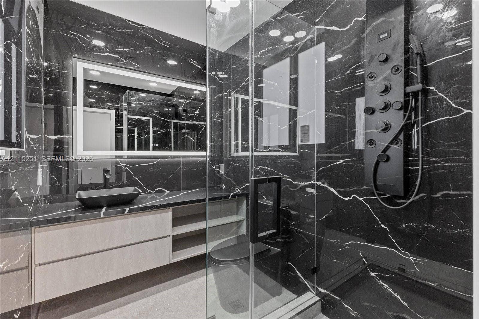 19511 NE 18th Place, Miami, FL 33179 Floating vanity with integrated shelving, dark-veined stone surfacing, rectangular vessel sink, LED-backlit mirror, and glass-enclosed shower featuring a multi-functional matte black shower panel