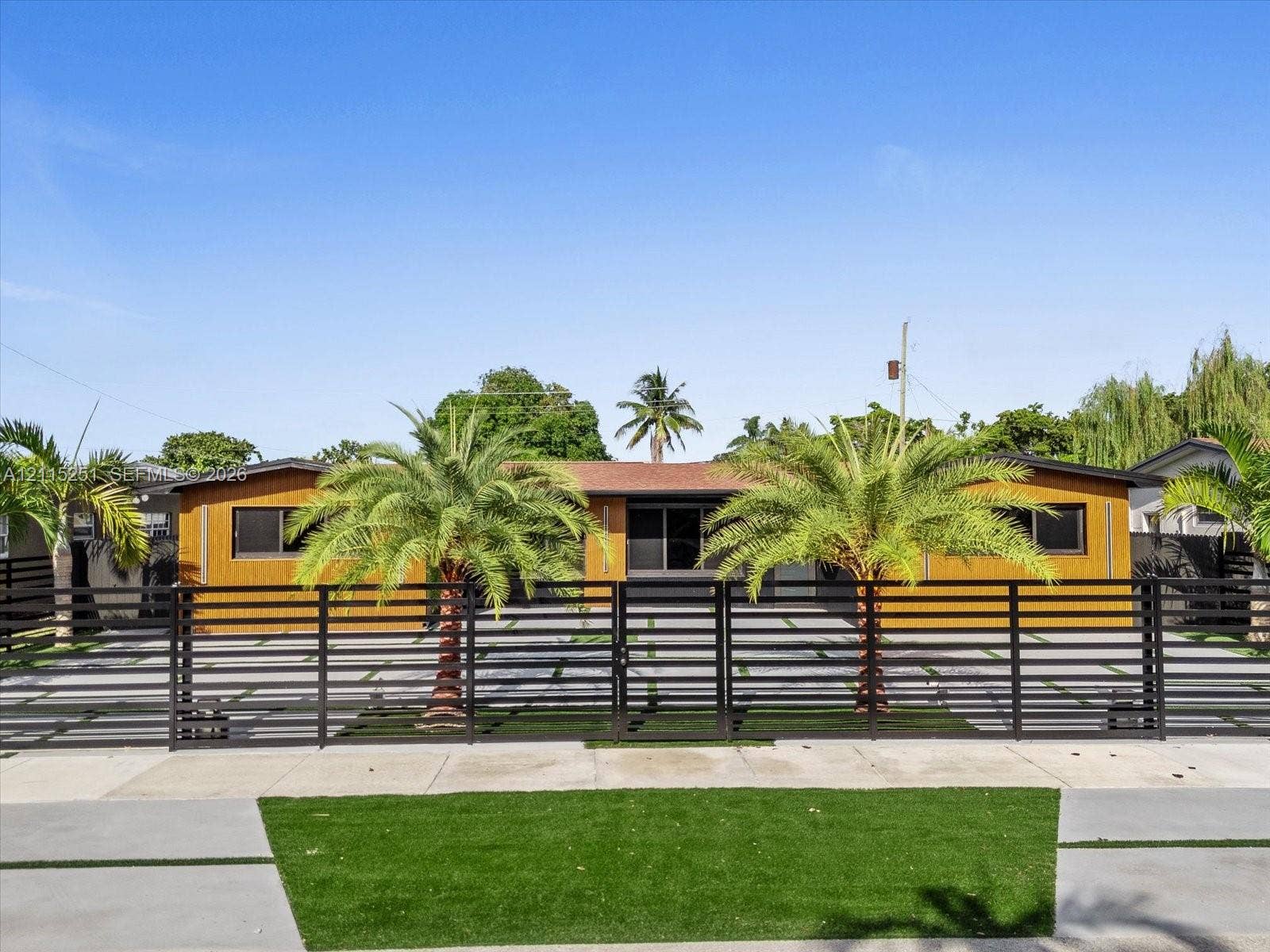 19511 NE 18th Place, Miami, FL 33179 Single-story exterior featuring wood-finish paneling, horizontal slat metal fencing, paved driveway with grass inserts, and integrated palm trees