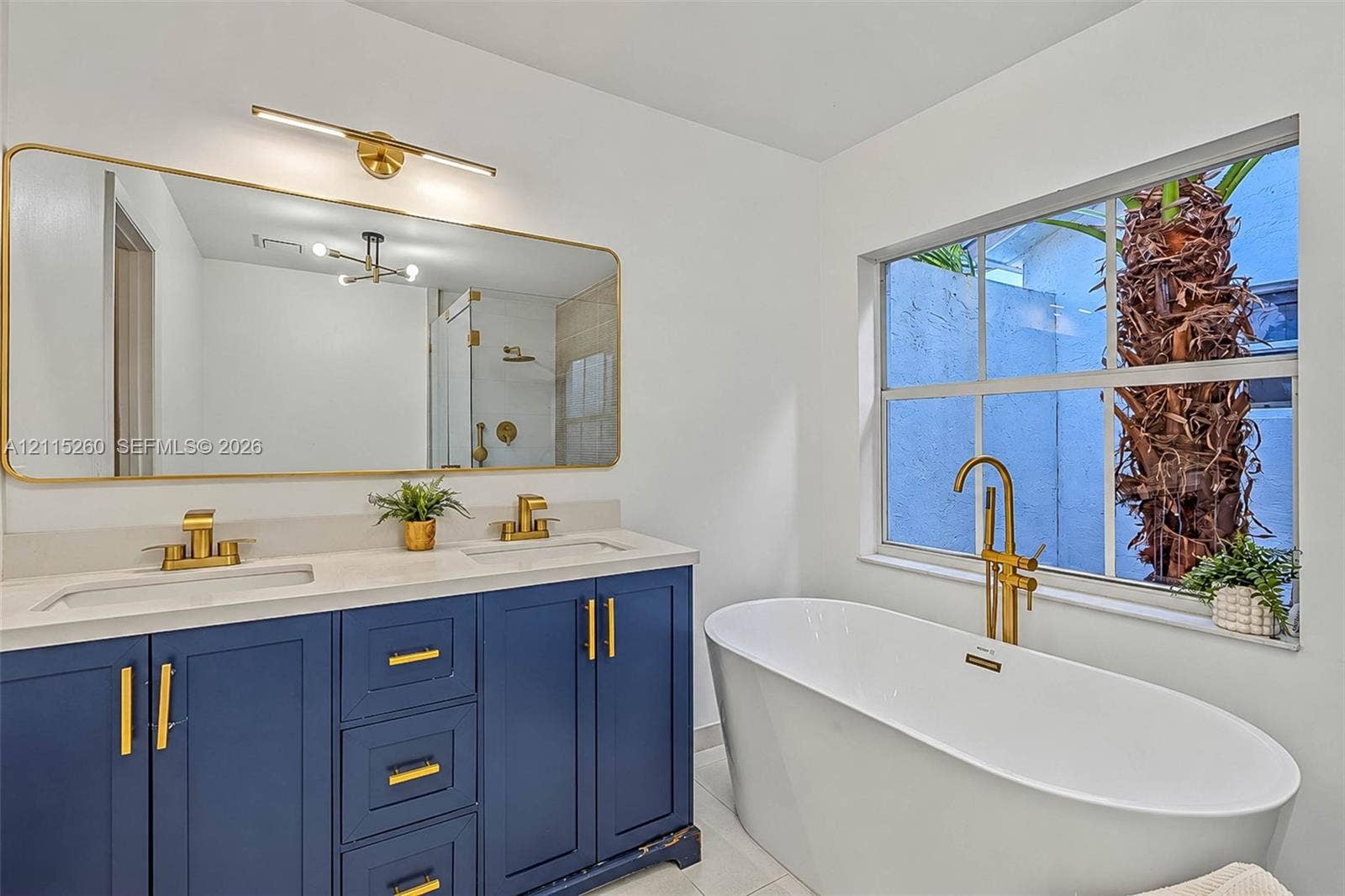 820 NW 132nd Court, Miami, FL 33182 Dual-sink vanity with navy blue cabinetry, gold-tone hardware, and a quartz countertop, paired with a freestanding soaking tub featuring a gold-tone floor-mounted faucet and an expansive gold-rimmed mirror