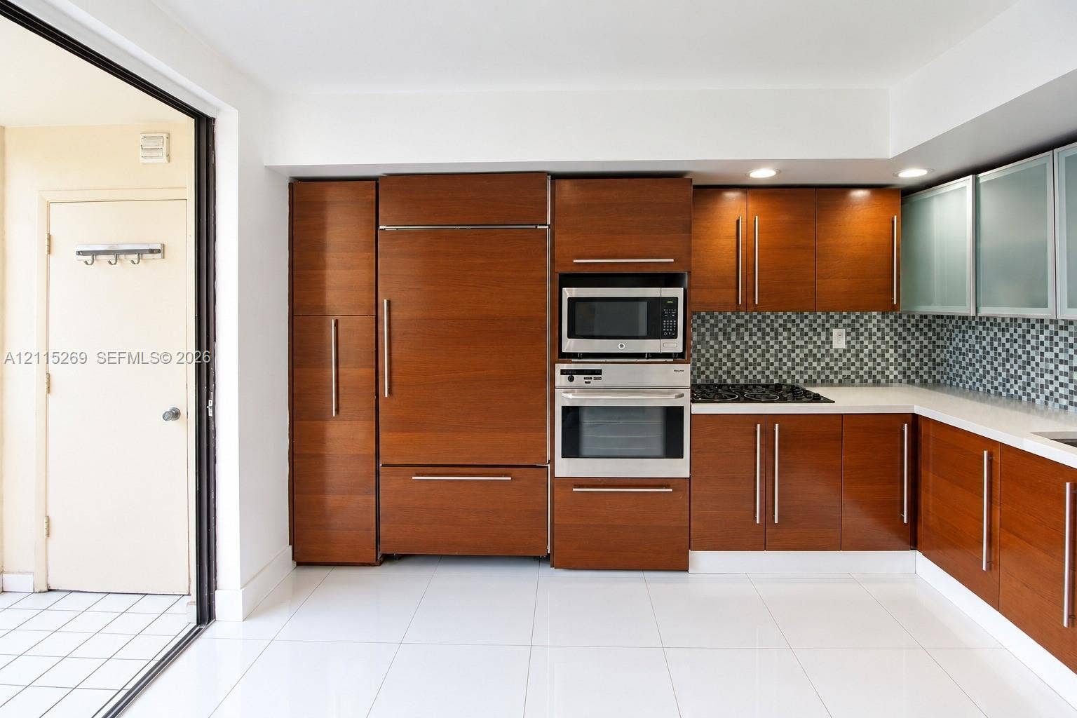 8615 NW 8th Street, #211, Miami, Miami, FL 33126 Kitchen featuring custom wood-finish cabinetry with integrated appliances, stainless steel wall-mounted oven and microwave, mosaic tile backsplash, solid-surface countertops, and large-format tile flooring