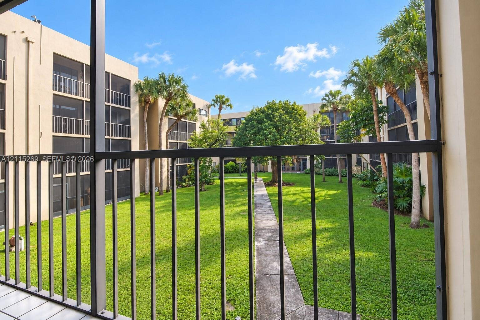 8615 NW 8th Street, #211, Miami, Miami, FL 33126 Screened balcony with metal railing overlooking a landscaped courtyard featuring a lawn, mature palm trees, and a paved walkway
