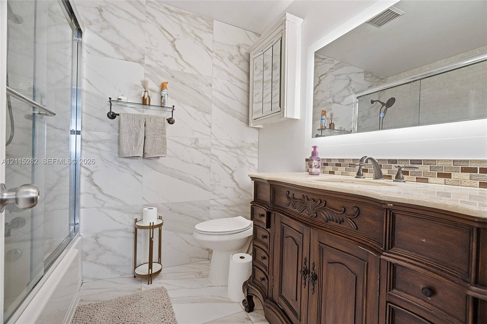 Bathroom featuring large-format stone-finish wall and floor tile, wood-finish vanity with stone countertop, undermount sink, and illuminated mirror