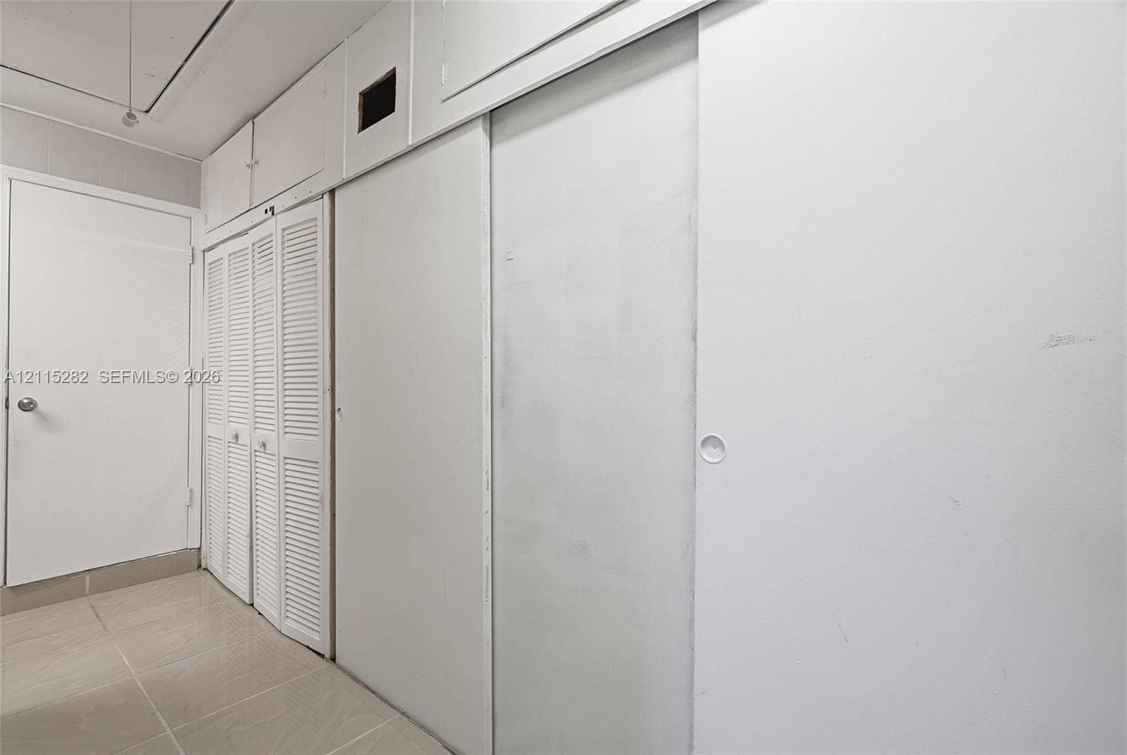 Wall-to-wall built-in storage featuring floor-to-ceiling smooth-panel sliding doors, overhead cabinet compartments, and louvered-style closet doors, complemented by ceramic tile flooring