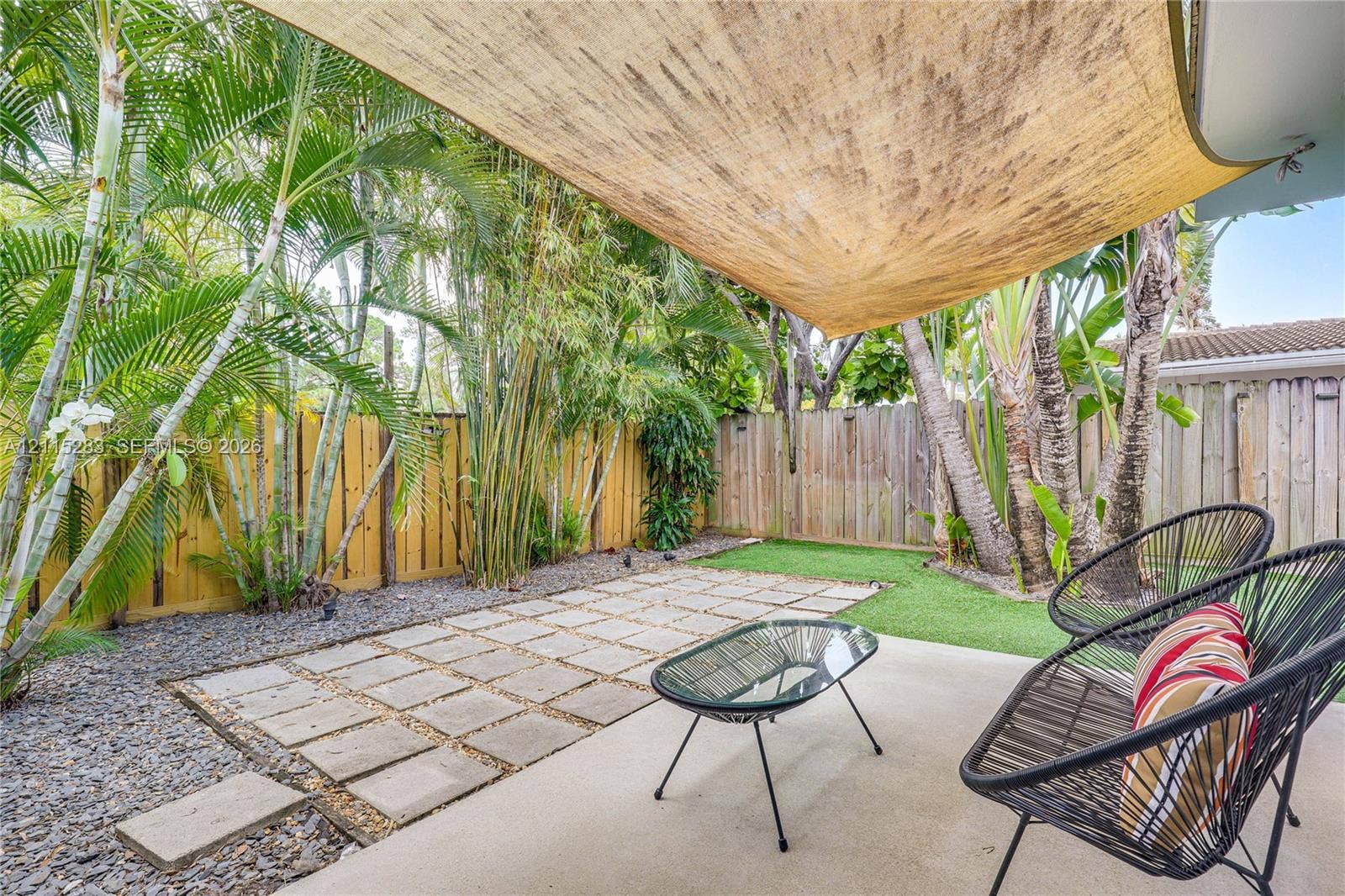 1619a NE 25th Street, #A, Wilton Manors, Wilton Manors, FL 33305 Covered concrete patio featuring a fabric shade sail, paver stone hardscaping, artificial turf, perimeter wood fencing, and mature palm landscaping