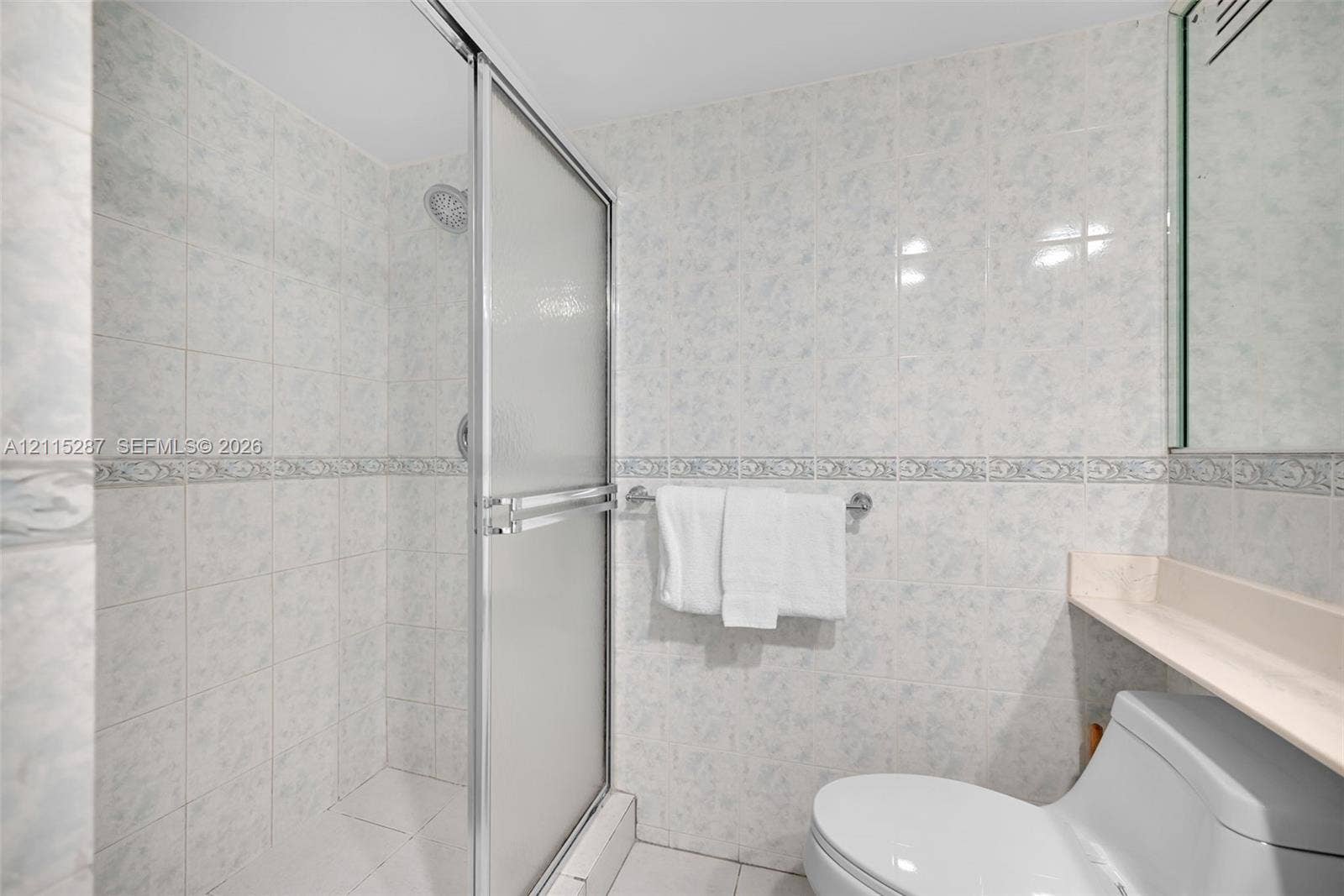 5225 Collins Avenue, #511, Miami Beach, FL 33140 Walk-in shower featuring a sliding glass enclosure, floor-to-ceiling ceramic tile wall finishes, decorative tile accent banding, and a wall-mounted vanity shelf