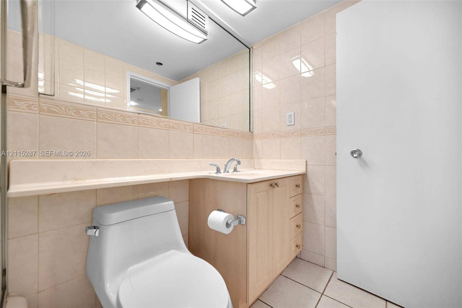 5225 Collins Avenue, #511, Miami Beach, FL 33140 Bathroom featuring a wood-finish vanity with an integrated countertop and sink, wall-to-wall mirror, tiled walls with decorative trim, and overhead task lighting