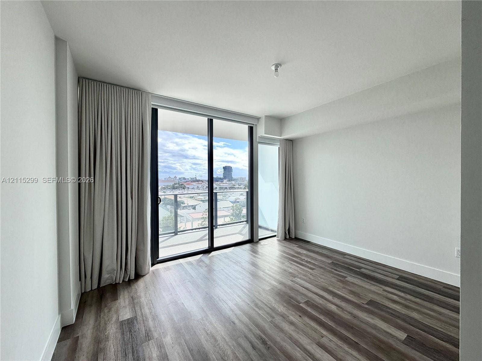 Floor-to-ceiling sliding glass door, private balcony with metal railing, wood-finish flooring, and expansive exterior urban scenery
