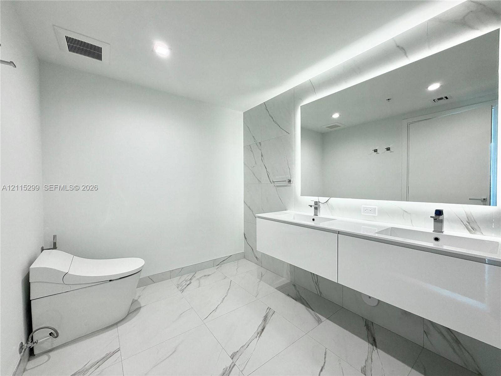 Modern floating double vanity featuring dual integrated sinks, large back-lit mirror, wall-mounted fixtures, and stone-finish floor and wall tiles