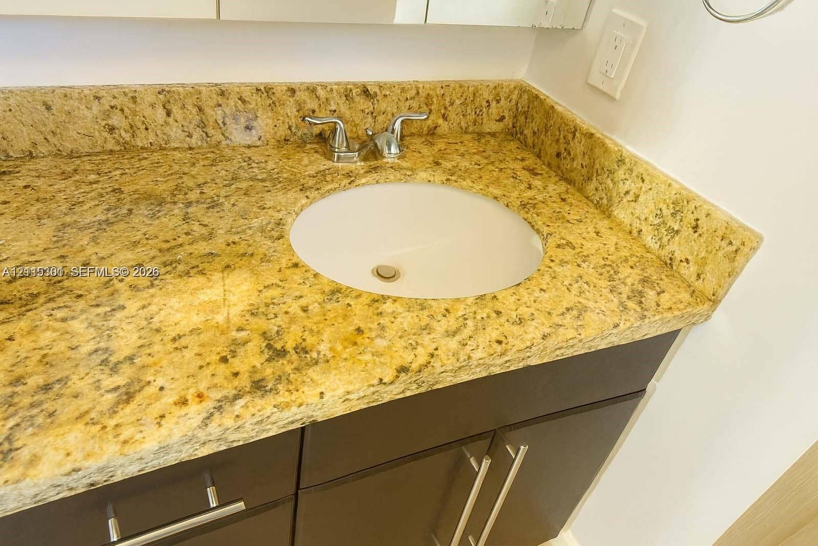 1630 Embassy Drive, #101, West Palm Beach, West Palm Beach, FL 33401 Vanity featuring a stone-composite countertop with an integrated undermount sink, chrome double-handle faucet, and dark-toned cabinetry with metallic hardware