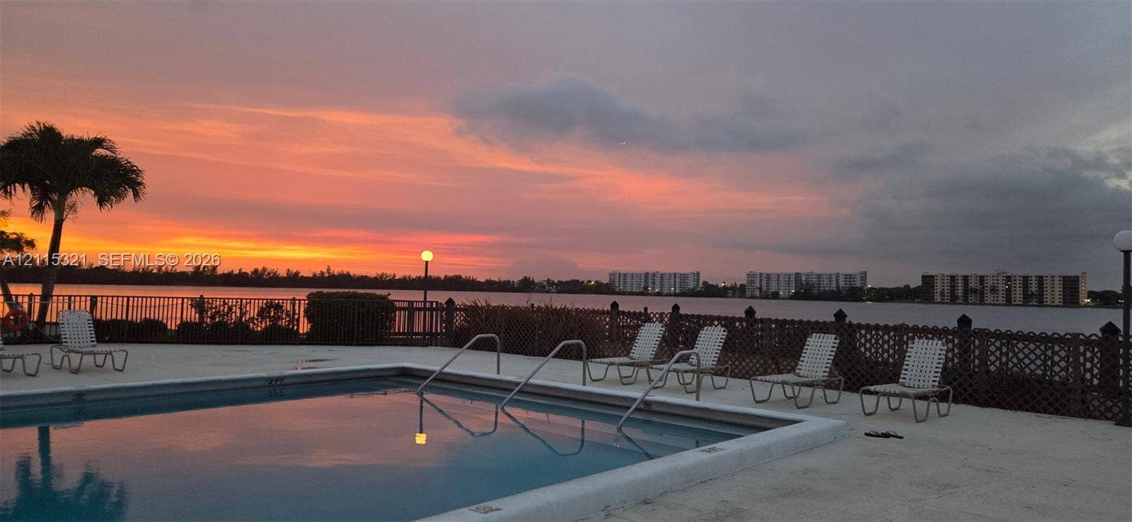 115 Lake Emerald Drive, #408, Oakland Park, FL 33309 Swimming pool with a concrete deck, metal perimeter fencing, and a waterfront setting against a sunset backdrop