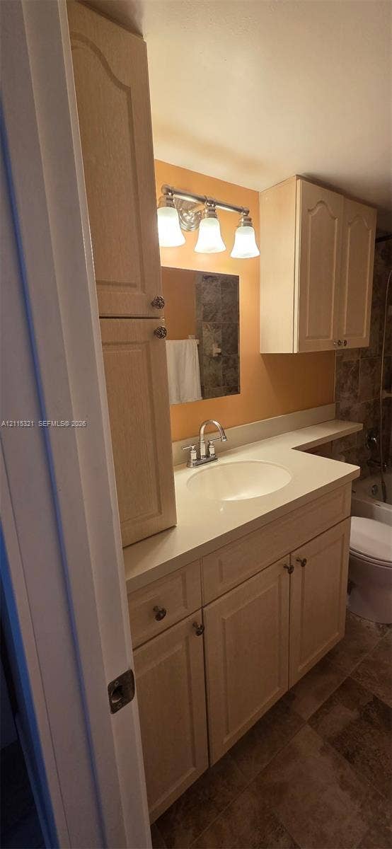 115 Lake Emerald Drive, #408, Oakland Park, FL 33309 Vanity featuring light wood-finish cabinetry, a solid-surface countertop, an integrated sink, a three-light wall fixture, and overhead storage cabinetry