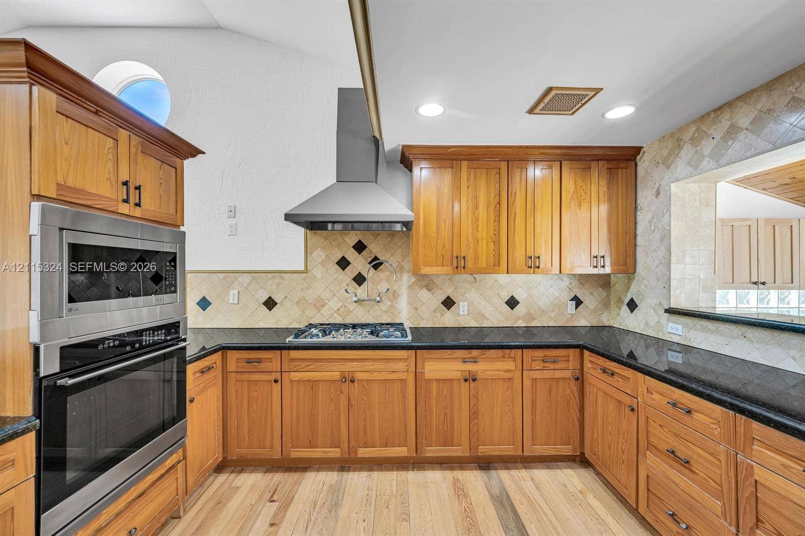 Kitchen featuring natural wood cabinetry, dark stone countertops, stainless steel wall-mounted oven and microwave, professional-style stainless steel range hood, and tile backsplash with decorative stone accents