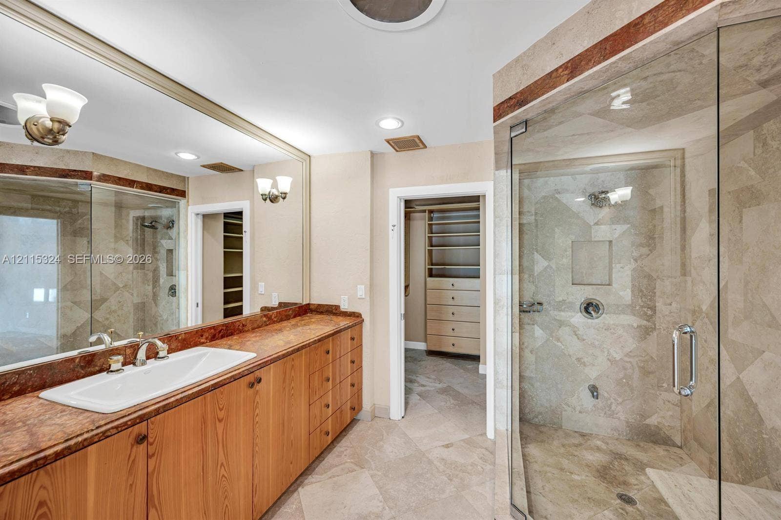 Bathroom featuring a stone-topped vanity with an integrated basin, expansive wall-mounted mirror, glass-enclosed shower with stone tile surround, and built-in closet cabinetry
