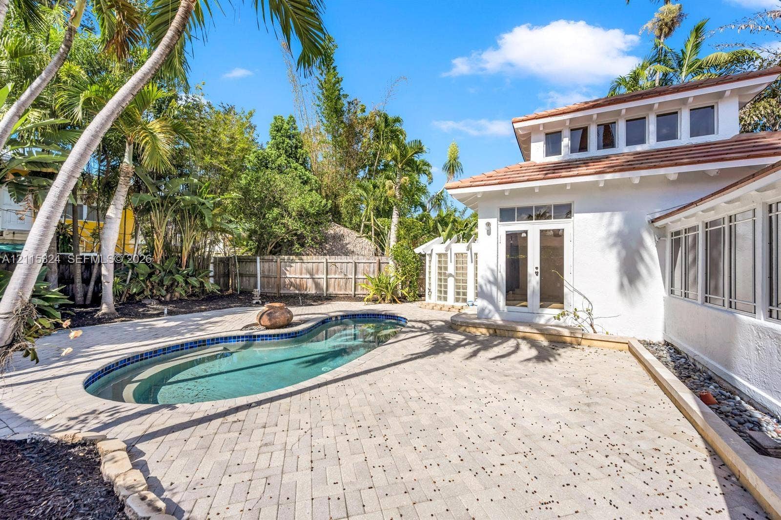 Free-form swimming pool with tile surround, expansive paver patio, white stucco exterior, tile roofing, and glass block accent walls