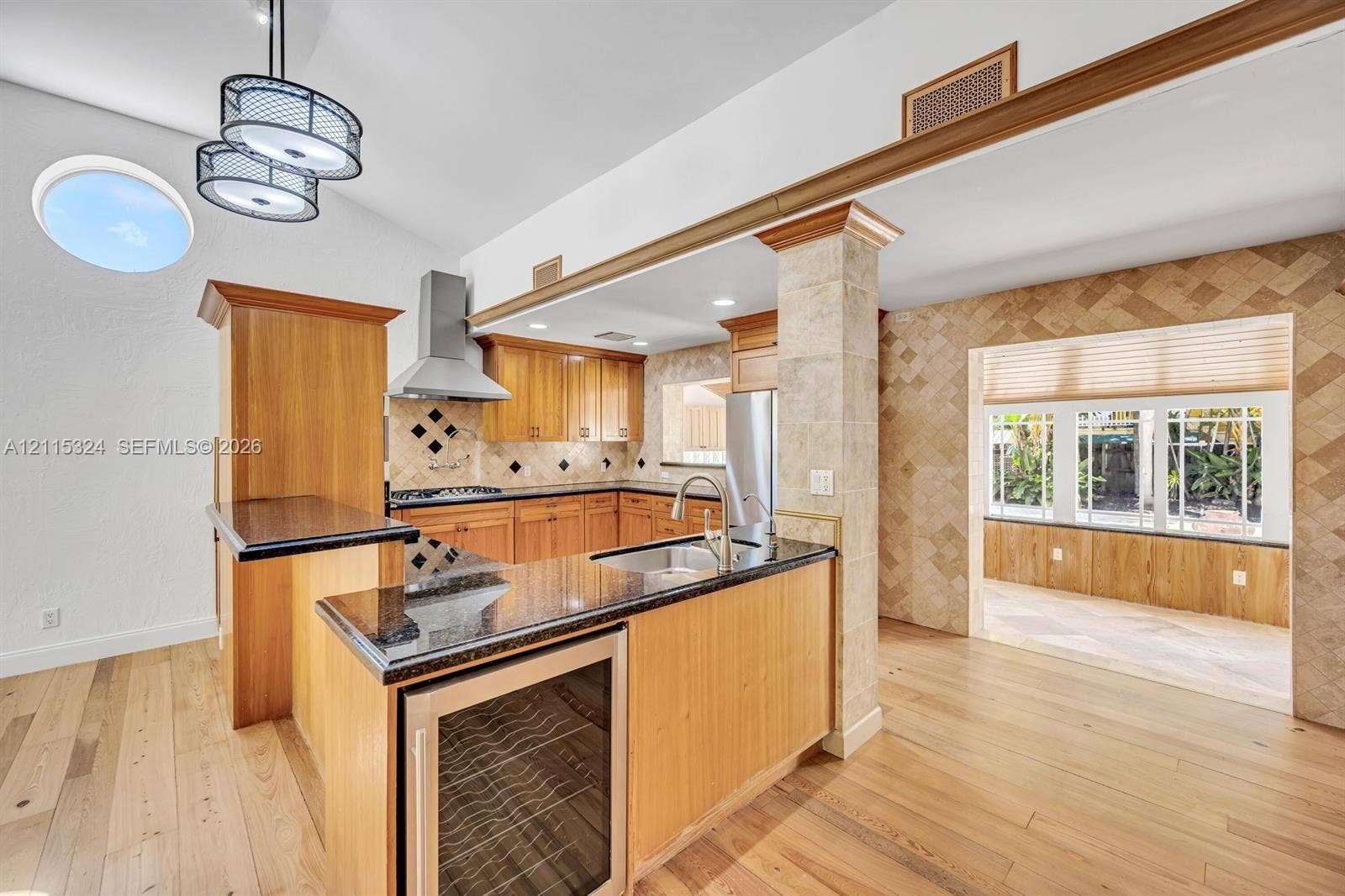 Kitchen featuring custom wood cabinetry, stone-topped island with integrated beverage refrigerator, stainless steel range hood, stone tile backsplash, and wood-finish flooring