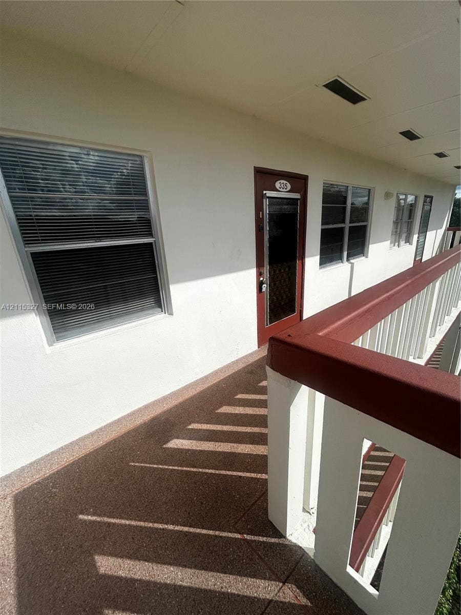 335 Brighton H, #H, Boca Raton, Boca Raton, FL 33434 Exterior walkway featuring textured stone-aggregate flooring, a white railing with a dark-tone handrail, and multiple windows with integrated blinds