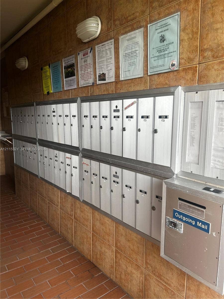 335 Brighton H, #H, Boca Raton, Boca Raton, FL 33434 Wall-mounted metallic mailbox station featuring individual locked compartments and an integrated outgoing mail slot, set against a ceramic-tiled wall with attached overhead lighting