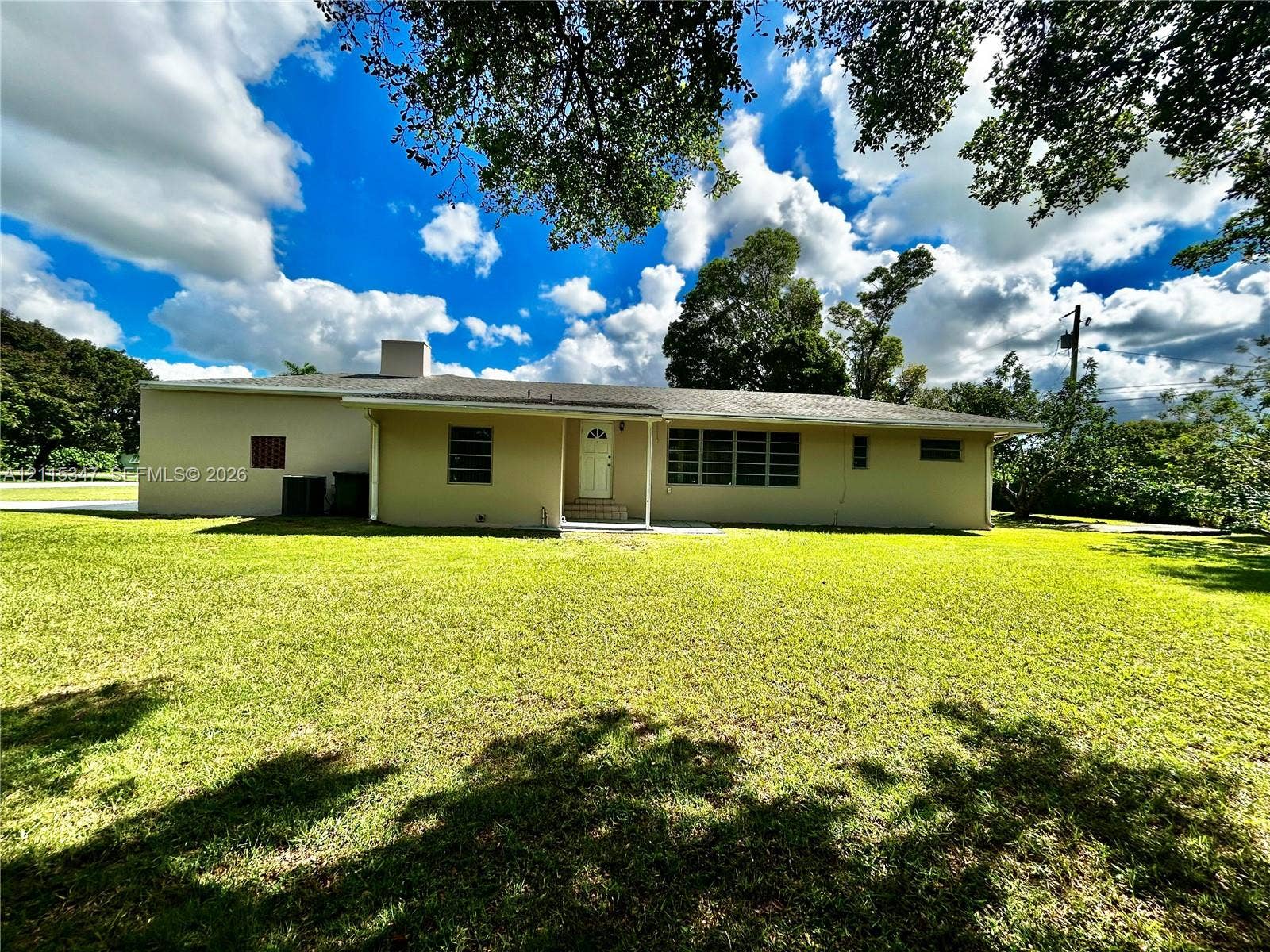 Single-story residence featuring a low-pitch roof, light-toned exterior finish, horizontal multi-pane windows, and a covered entryway set upon an expansive lawn with mature shade trees