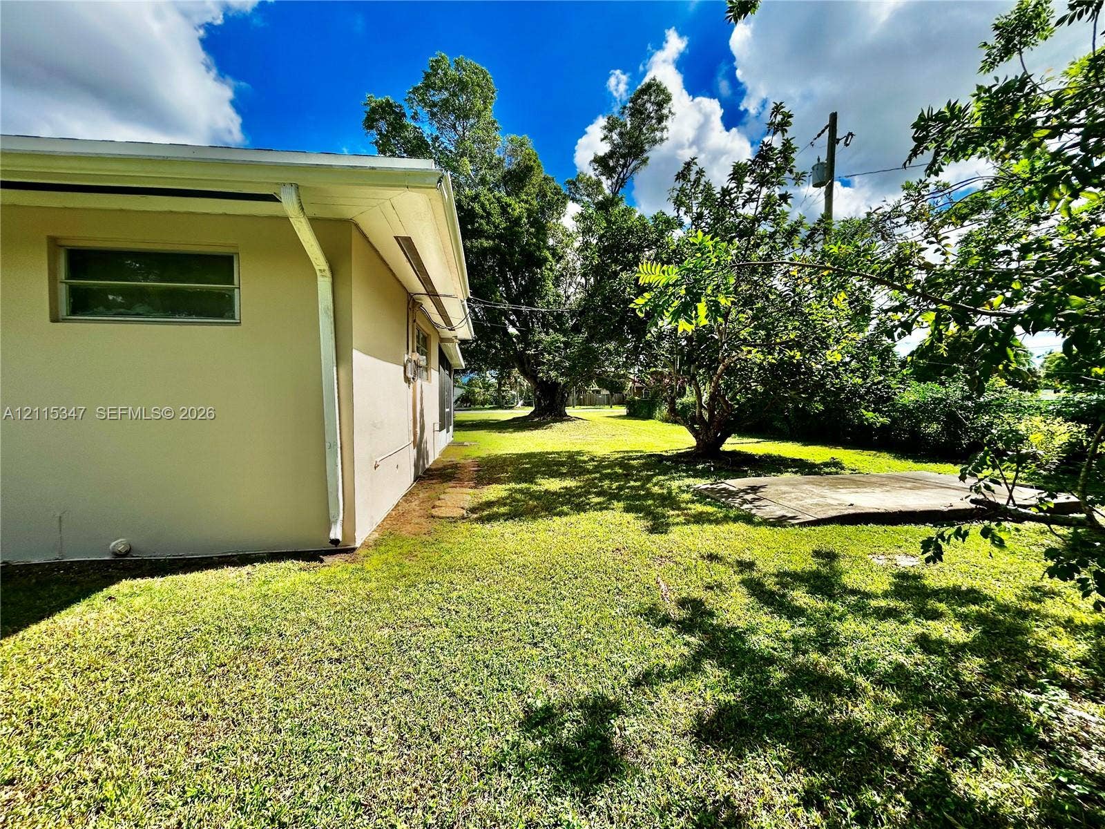 Exterior side yard featuring stucco siding, a concrete slab, mature trees, and grass lawn