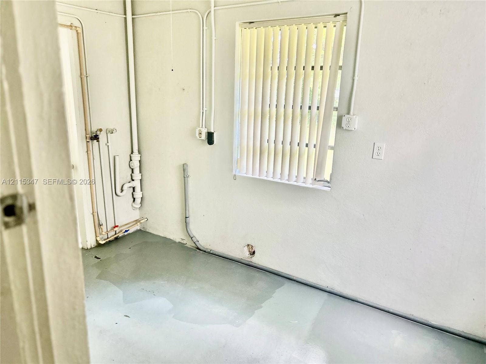 Utility area featuring exposed plumbing lines, a dedicated drain pipe, a window with vertical blinds, and a concrete floor finish