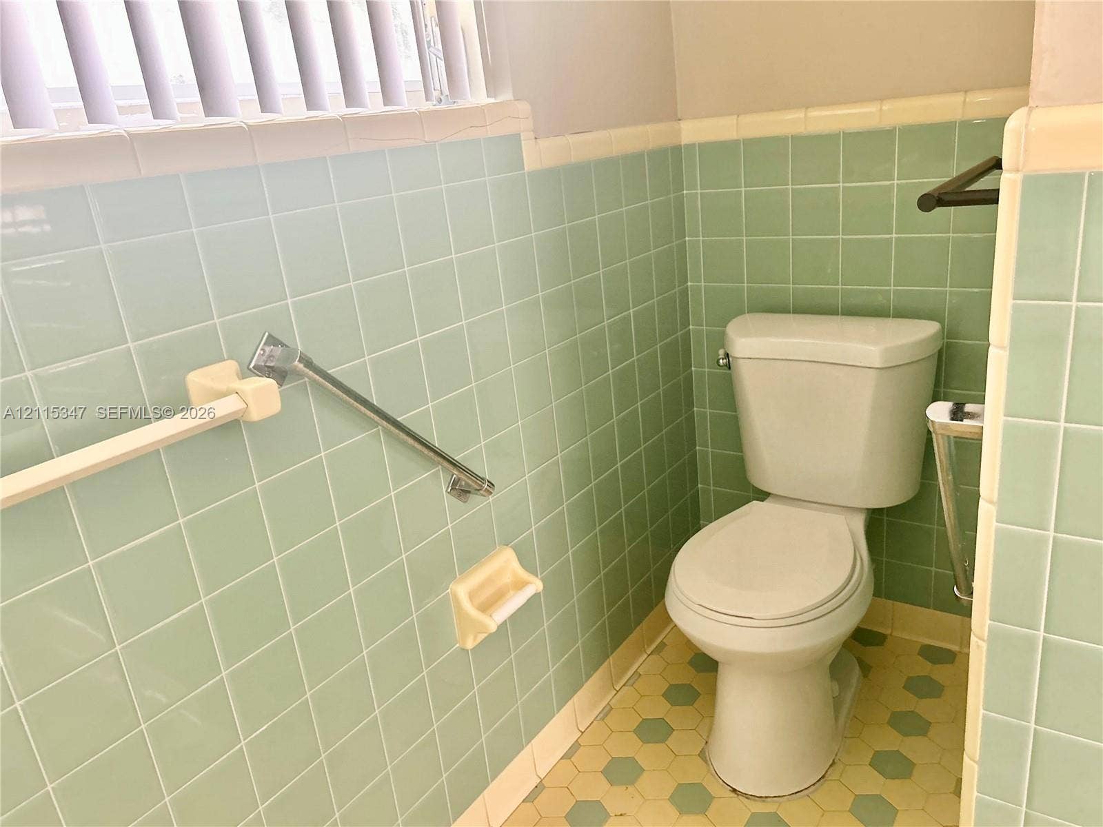 Restroom featuring light green square wall tiling, cream-colored trim, hexagon-patterned flooring, a two-piece toilet, and metallic wall-mounted hardware