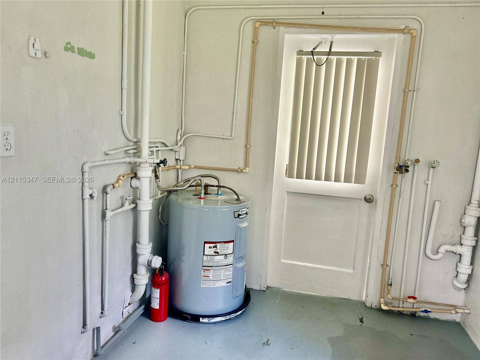 Utility area featuring a water heater, exposed plumbing lines, and an exterior door with vertical blinds