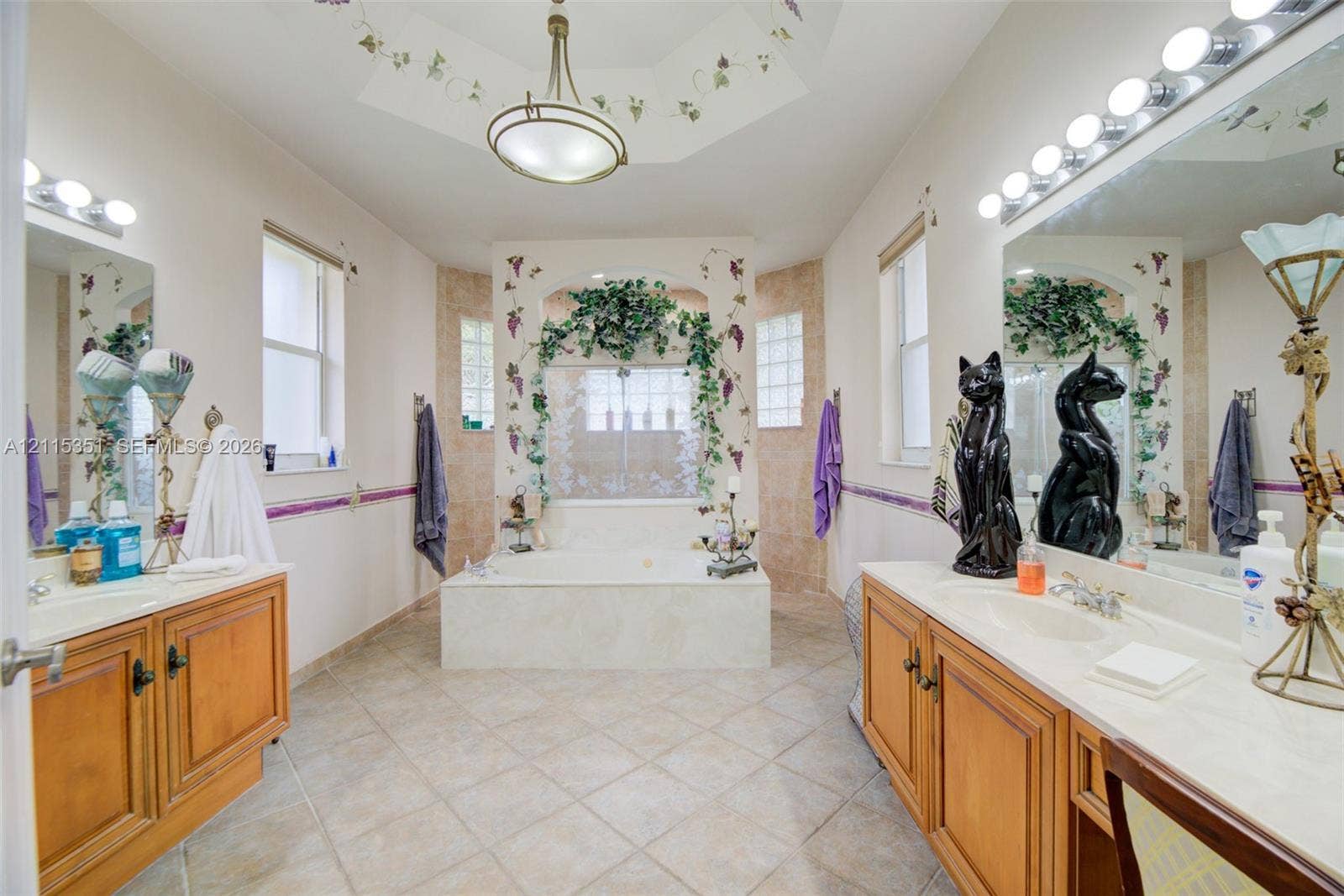 13845 NW 20th Street, Pembroke Pines, FL 33028 Bathroom featuring dual wood-finish vanities with solid-surface countertops, a central jetted tub with tiled surround, glass block windows, and a coffered ceiling with a central light fixture