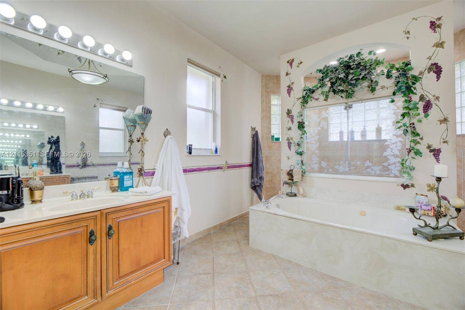 13845 NW 20th Street, Pembroke Pines, FL 33028 Bathroom featuring a wood-finish vanity with a molded countertop, expansive wall-mounted mirror with integrated lighting, oversized soaking tub with an arched glass-block surround, and tiled flooring