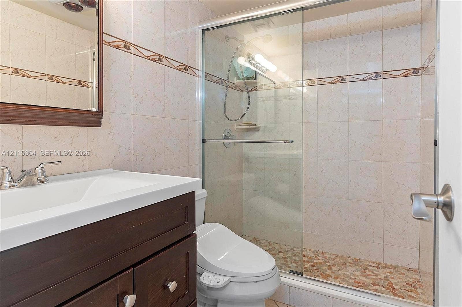 1980 S Ocean Drive, #20A, Hallandale Beach, Hallandale Beach, FL 33009 Bathroom featuring a dark wood-finish vanity with a white integrated basin, tile wall cladding with a decorative border, a glass-enclosed shower with pebble-stone flooring, and a wall-mounted mirror with a dark wood frame