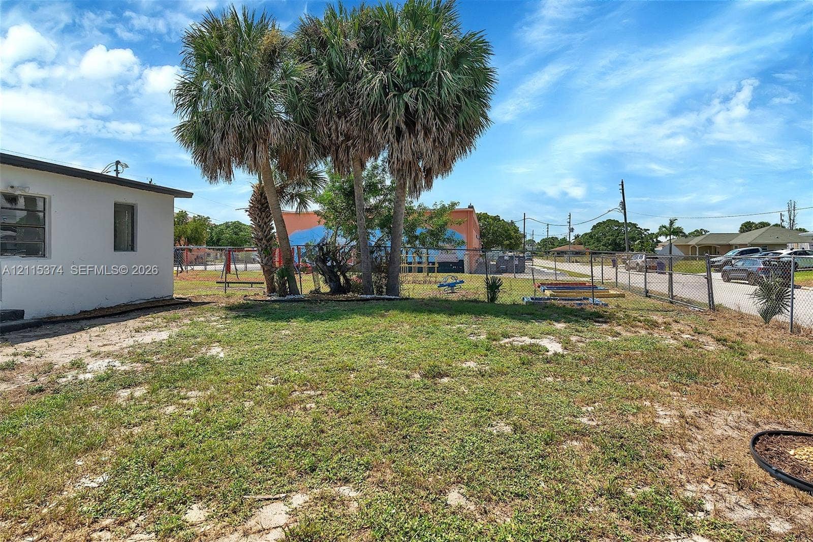 Exterior yard space featuring mature palm trees, perimeter chain-link fencing, and a combination of grass and sand landscaping