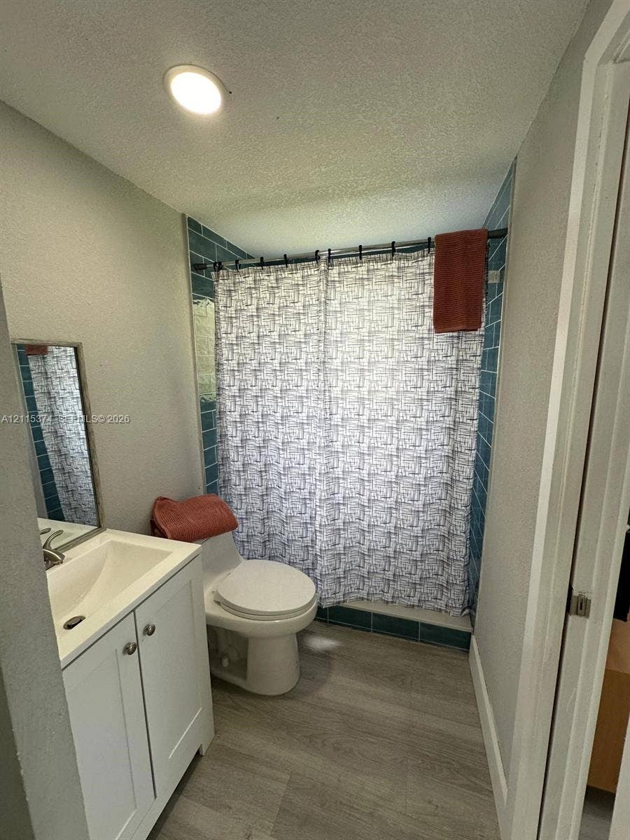 Bathroom featuring wood-finish flooring, white vanity cabinet with integrated sink, rectangular mirror, and a tiled shower surround