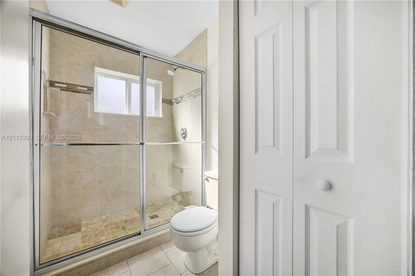 Shower enclosure with sliding glass doors, neutral tile wall surround with decorative inlay, inset window, and classic white paneled closet doors
