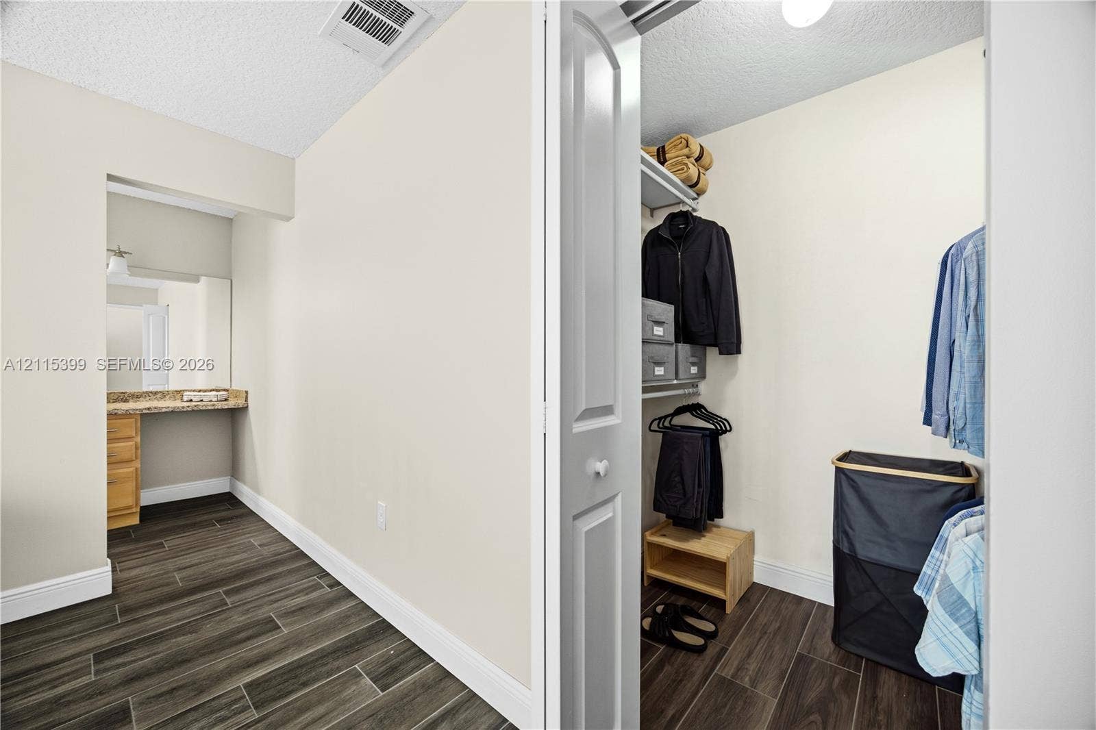 Walk-in closet featuring built-in wire shelving and wood-finish flooring, complemented by a vanity area with wood cabinetry and stone-finish countertop