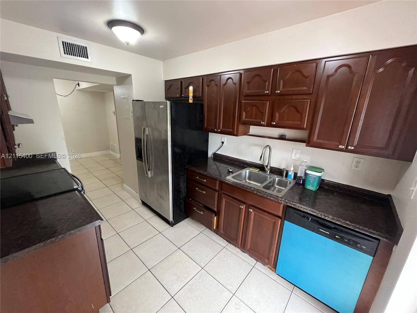 Kitchen featuring dark wood-finish cabinetry, stone-surface countertops, stainless steel refrigerator, dual-basin sink with arched faucet, and tiled flooring