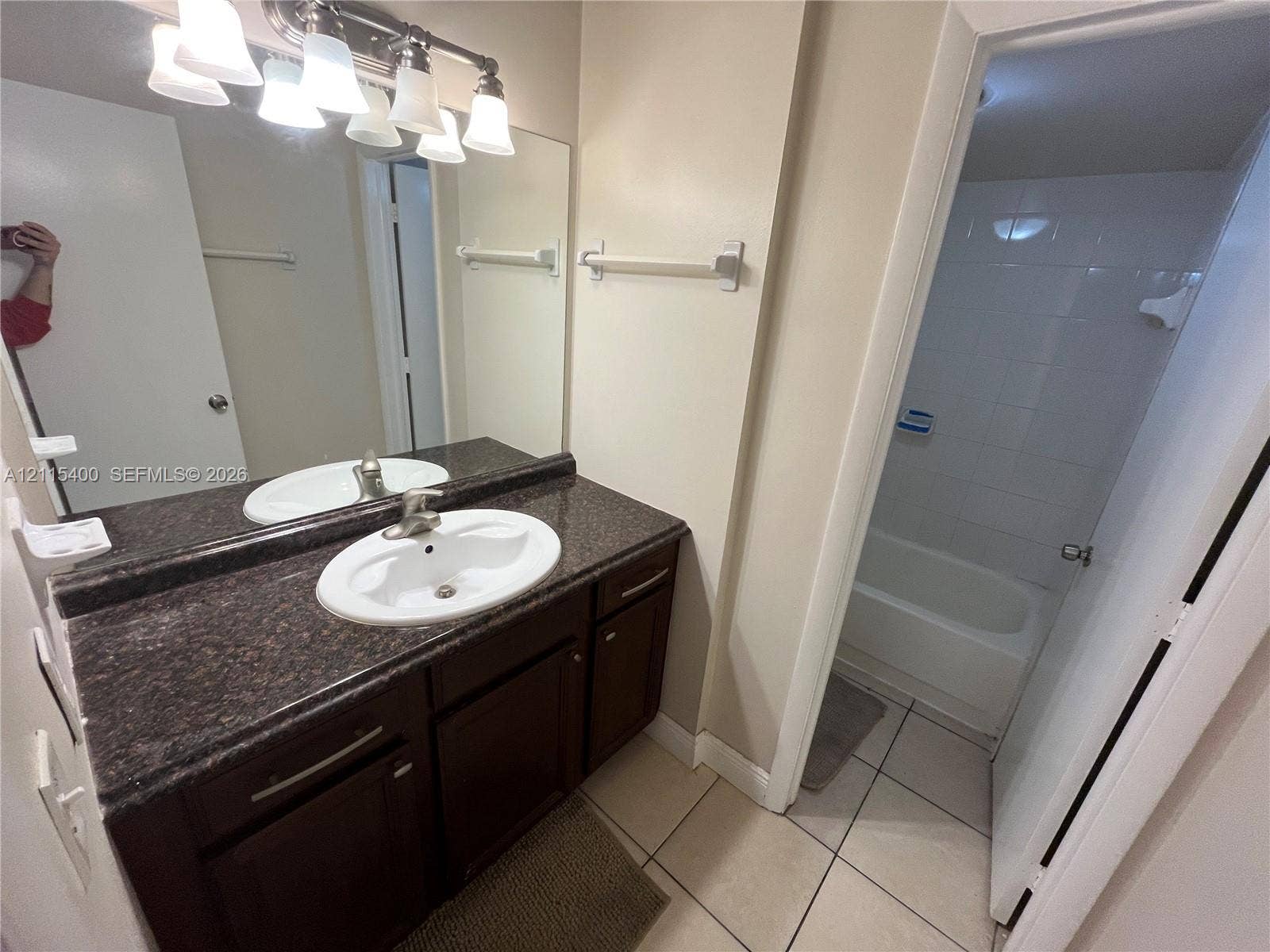 Bathroom vanity featuring dark-toned cabinetry, a stone-finish countertop with an integrated basin, a full-width mirror, and a wall-mounted light fixture, paired with a shower-tub combination featuring wall-surround tiling and tile flooring