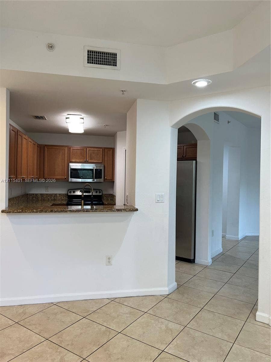 11503 NW 89th Street, #104, Doral, Doral, FL 33178 Kitchen featuring wood cabinetry, stone-surface countertops, stainless steel appliances, and tile flooring, highlighted by an arched architectural opening