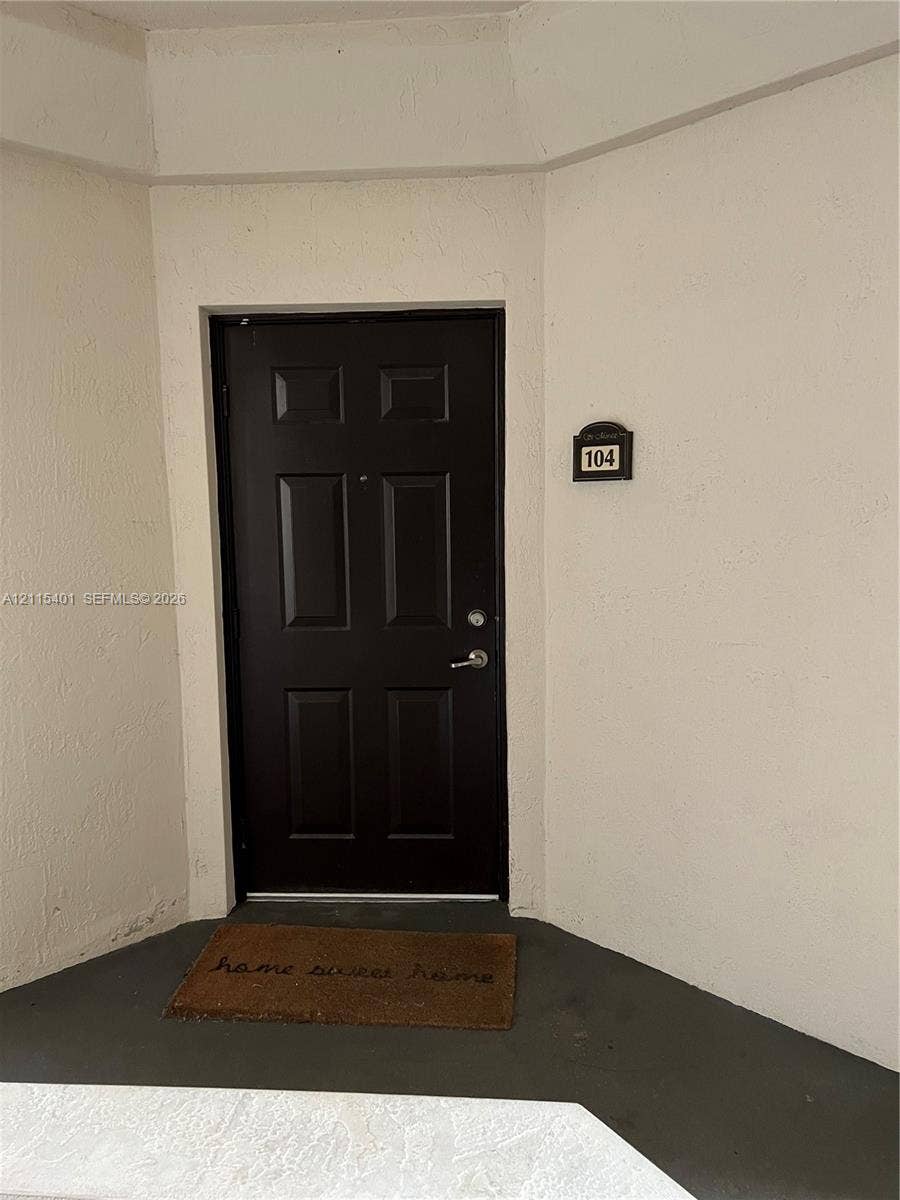 11503 NW 89th Street, #104, Doral, Doral, FL 33178 Dark-toned six-panel entry door featuring a metal lever handle and integrated deadbolt, set against a neutral-toned stucco exterior wall