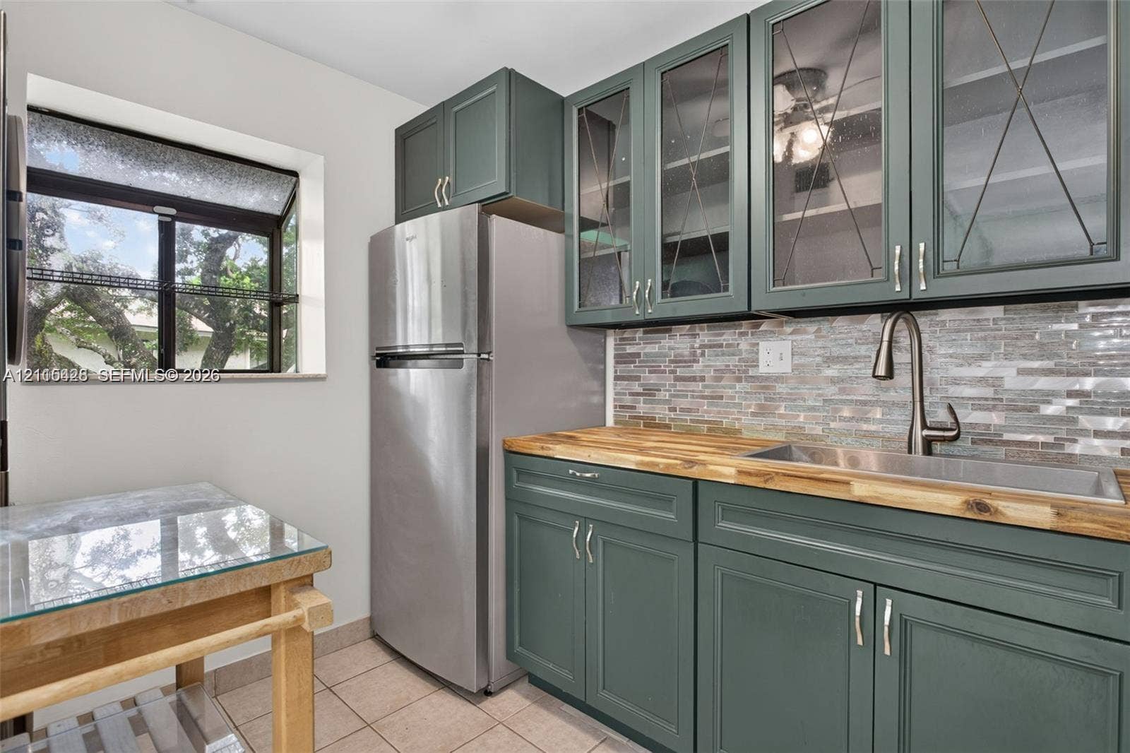 503 Gardens Drive, #201, Pompano Beach, Pompano Beach, FL 33069 Kitchen featuring blue-green shaker cabinetry with glass-front uppers, butcher block countertops, stainless steel undermount sink with gooseneck faucet, and mosaic tile backsplash