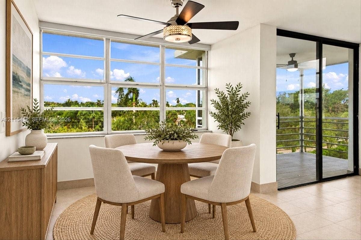 Sun-filled room featuring large, multi-pane windows, neutral tile flooring, a decorative ceiling fan with an integrated light fixture, and a sliding glass door with a metal-railed balcony
