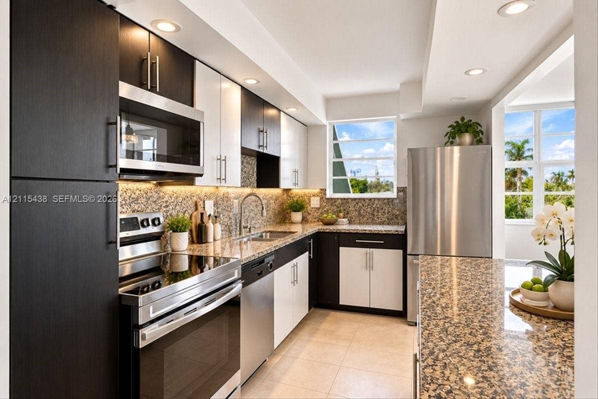 Two-tone cabinetry with contrasting upper and lower finishes, stone countertops, stainless steel appliances, recessed ceiling lighting, and tile flooring
