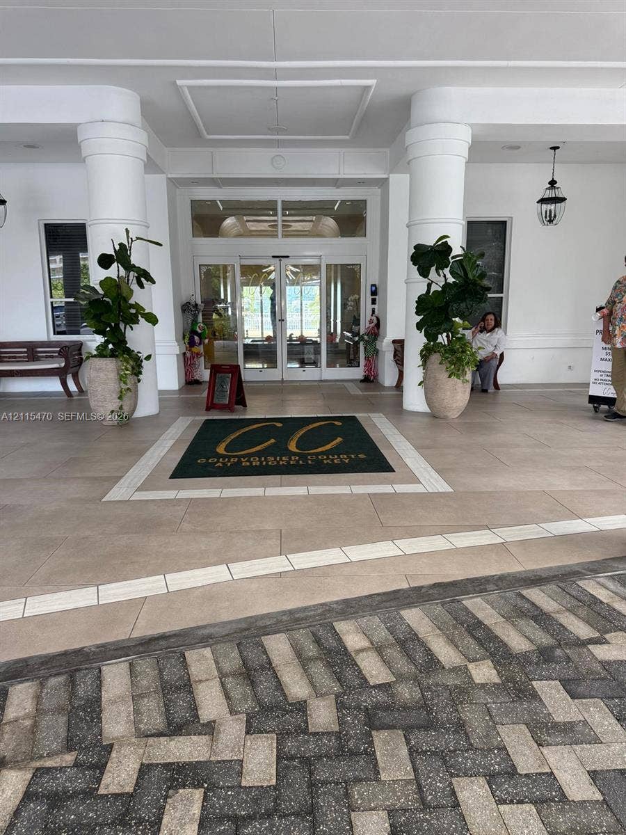 Covered building entrance featuring large structural columns, oversized tile flooring with a central inlay, glass-paned double entry doors, and wall-mounted exterior lanterns