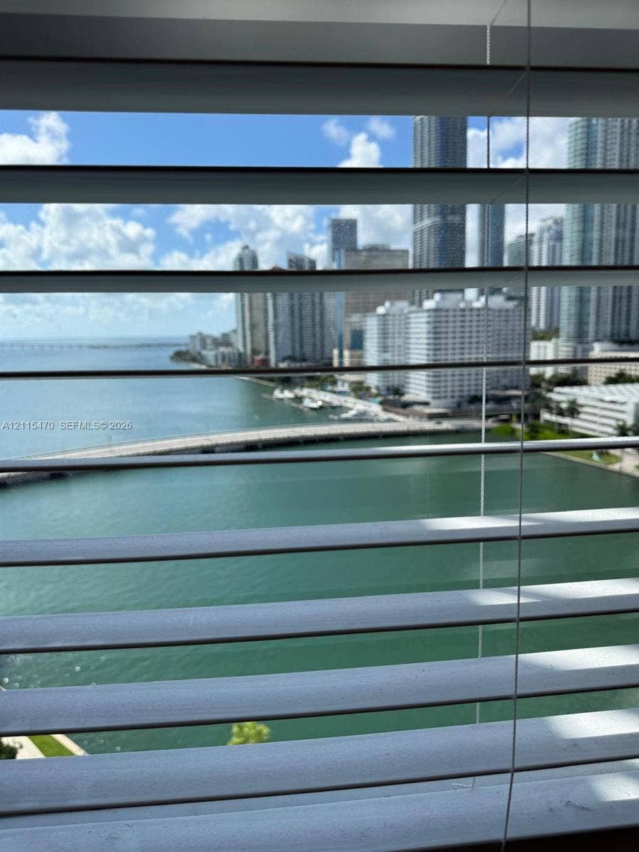 Horizontal window blinds framing a scene of the urban skyline, coastal waters, and a bridge